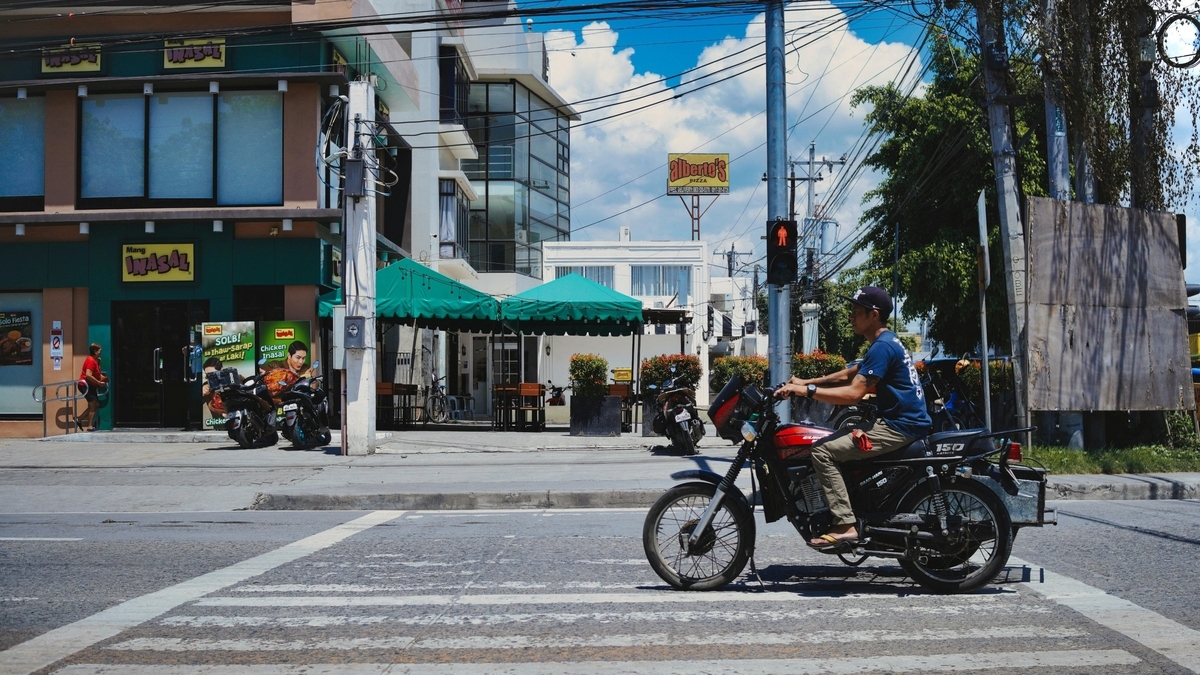 青空の下、赤と黒のバイクに乗った男性が横断歩道を渡る様子。背景には緑色の看板が特徴的な飲食店や白い建物、電柱が並び、フィリピンの街角の日常的な風景が広がっている。