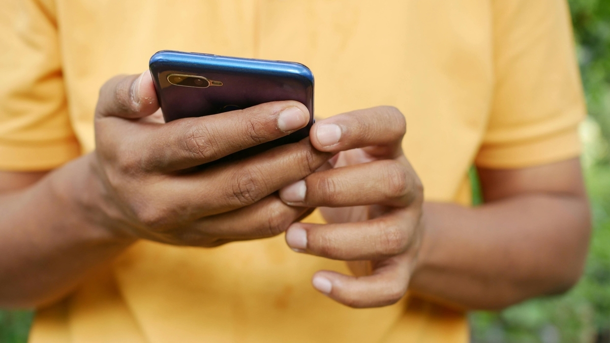 黄色の服を着た人物が、深い青色のスマートフォンを両手で持ち、画面を操作している手元のクローズアップ。指先がデバイスに触れ、屋外の光の中で使用している様子が描写されている
