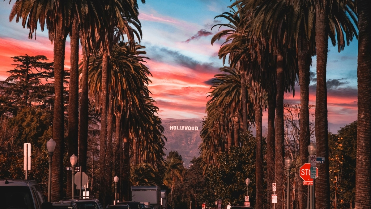 夕焼けの赤い空を背景に、両脇に背の高いヤシの木が並ぶ道路。正面の山の上には「HOLLYWOOD」の白い文字看板が見え、手前には道路標識や車のテールランプが写っている。