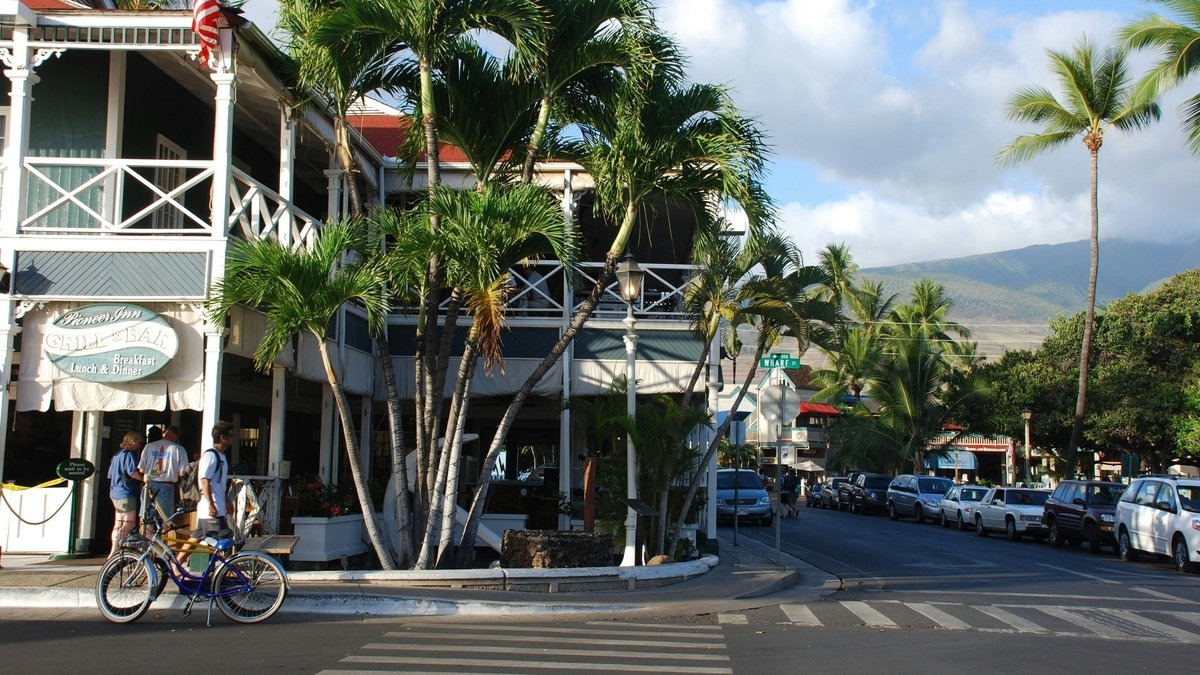 ヤシの木が立ち並ぶ通り沿いにある「Pioneer Inn GRILL & BAR」という看板を掲げた洋風の建物の前を、自転車に乗った人が通り過ぎる様子
