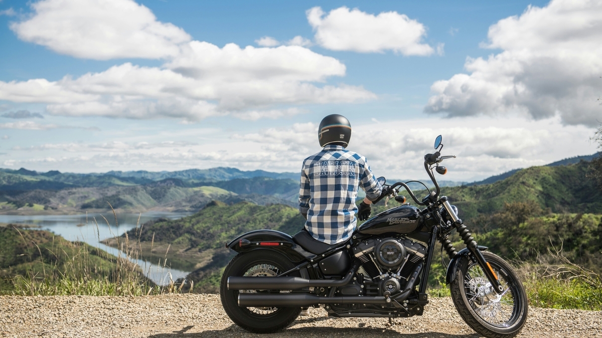 青空と白い雲、緑の山々と湖をバックに、黒いバイクにまたがり後ろ姿を見せるチェック柄シャツの人物