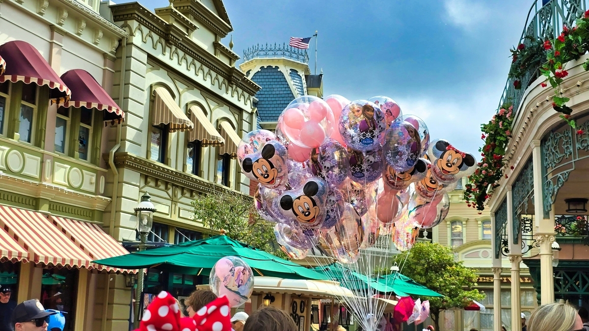 ディズニーランドのメインストリートUSA。クラシカルな建物と緑の日よけ傘を背景に、ミッキーマウスやミニーマウス、アナと雪の女王のキャラクターが描かれた透明なバルーンの束が浮かんでいる