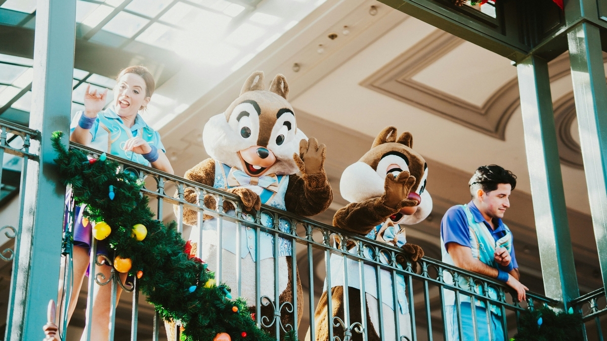 クリスマスのガーランドで飾られた建物のバルコニーから、手を振るチップとデールのキャラクター。両隣にはブルーのシャツを着たパークのキャストが並び、来園者に向かって笑顔を見せている