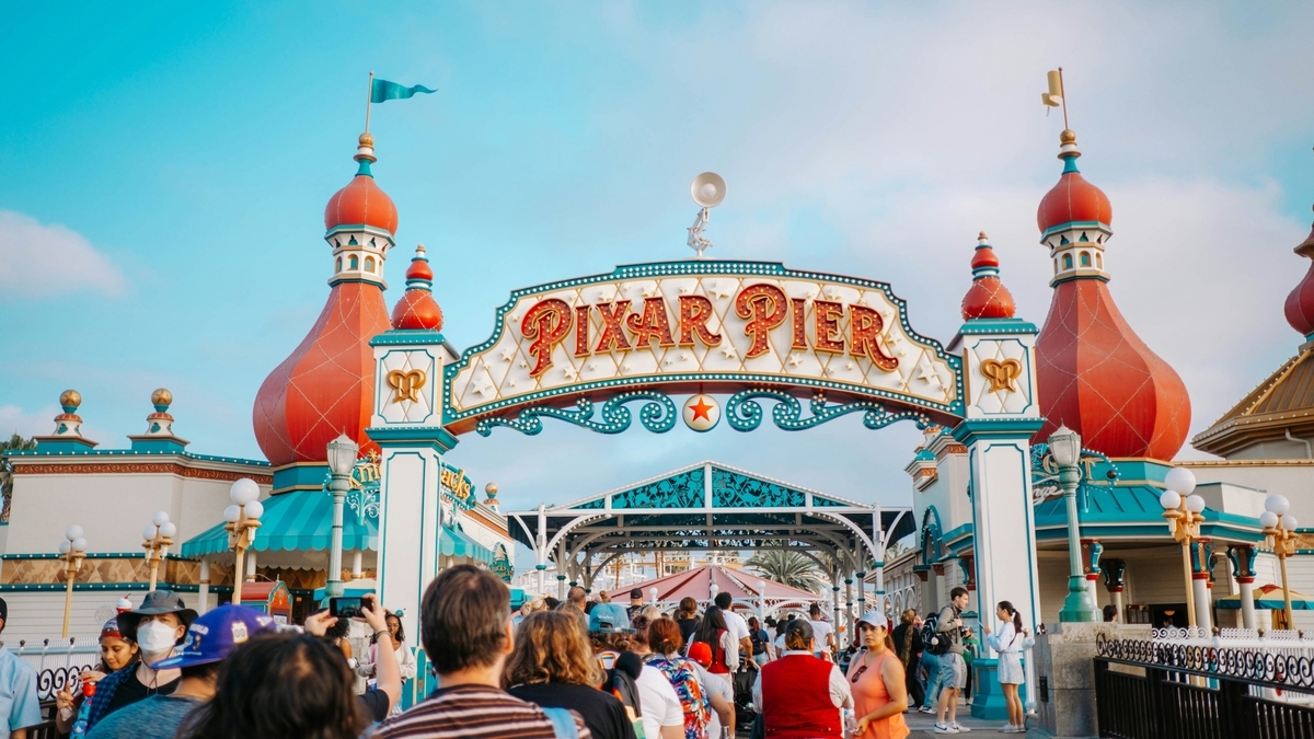 青空を背景に立つ、白とエメラルドグリーンを基調とした「PIXAR PIER（ピクサー・ピア）」のエントランスアーチ。アーチの先へ向かってマスクを着用した人々を含む多くの来園者が進んでいる。