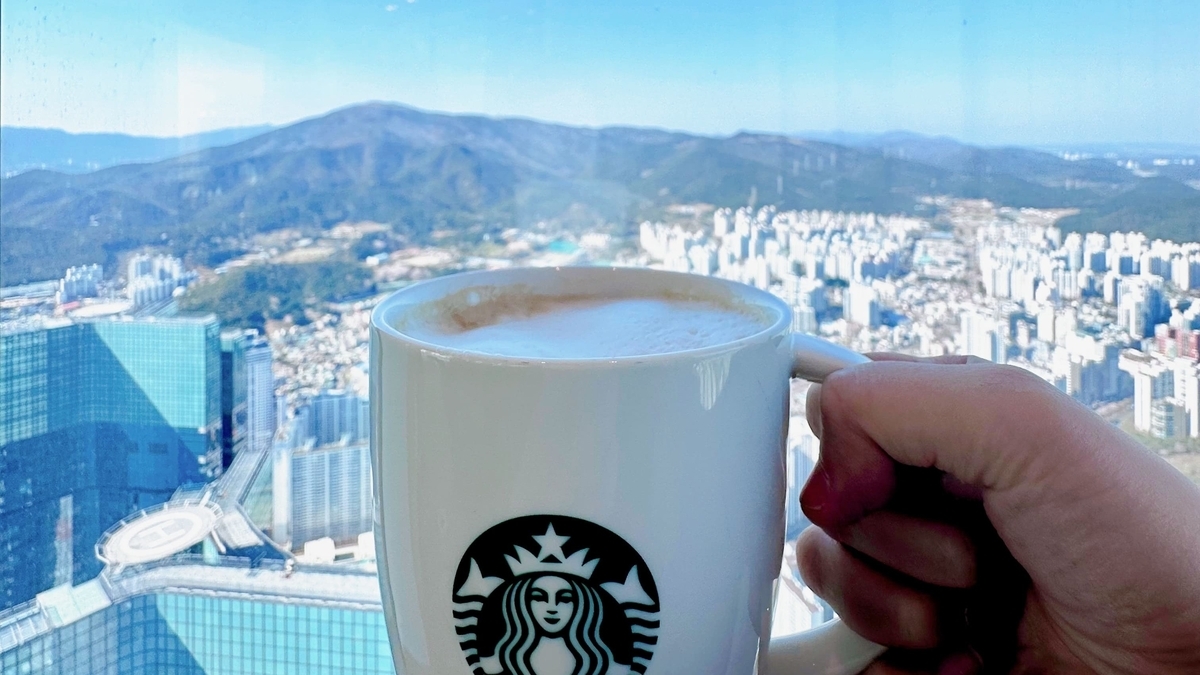 高層ビルの窓越しに広がる広大な街並みと山々を背景に、ロゴ入りの白いマグカップを持った手