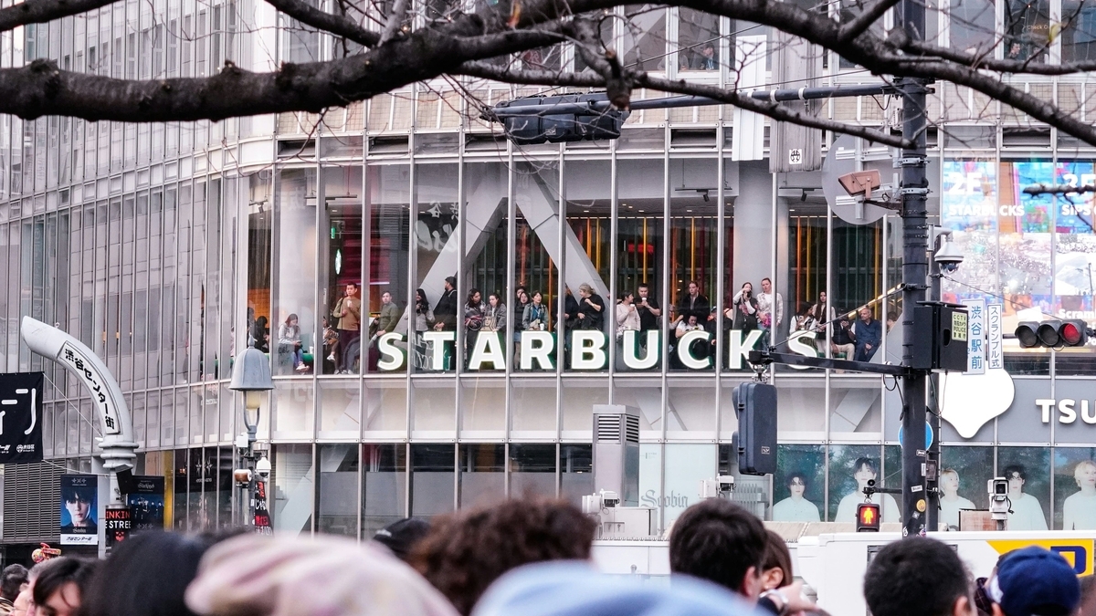 賑やかな通り沿いにあるガラス張りの近代的なビルの2階部分に、大きな「STARBUCKS」の文字が設置され、窓際に並ぶ多くの人々が外を眺めている様子