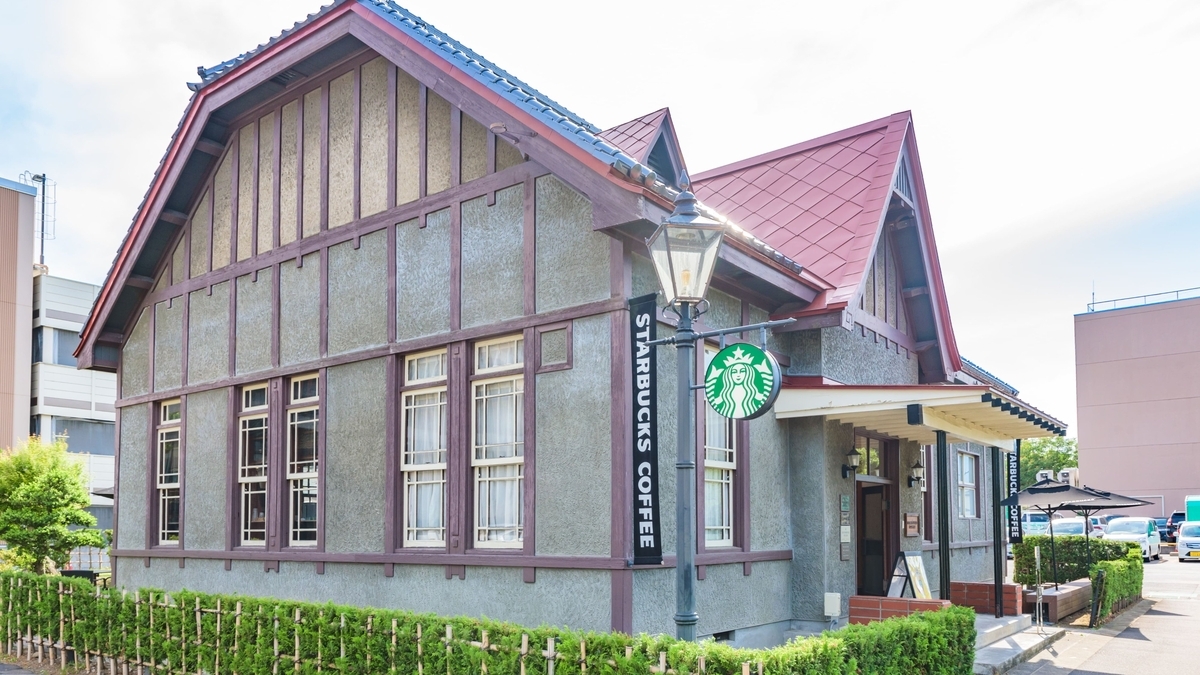 ハーフティンバー様式のような茶色い柱とグレーの壁の洋館で、緑のロゴ入りの街灯が前に立つ店舗外観