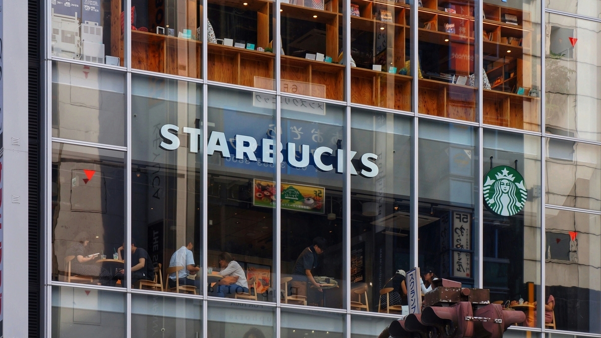 ガラスファサードのビルに「STARBUCKS」の白い文字と丸いロゴマークが掲げられ、ガラス越しに店内で過ごす人々の姿や木製の棚が見える外観