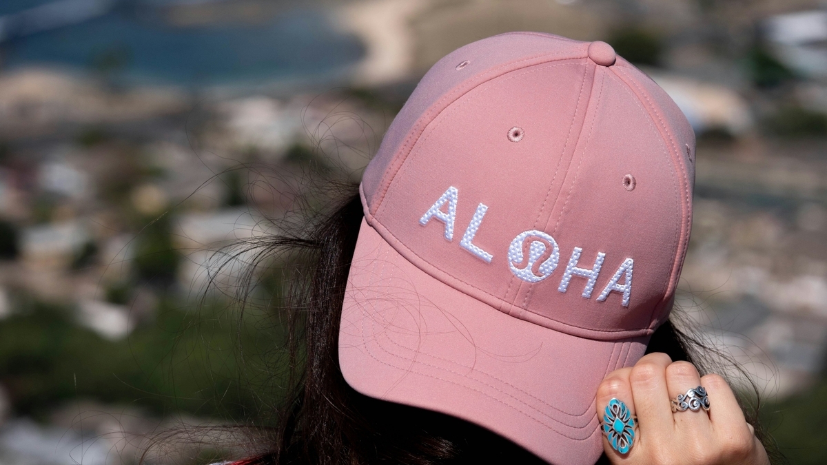 海と街並みを見下ろす景色を背景に、白文字でALOHAと刺繍されたピンク色のキャップをかぶり、指輪をはめた手を添える人物