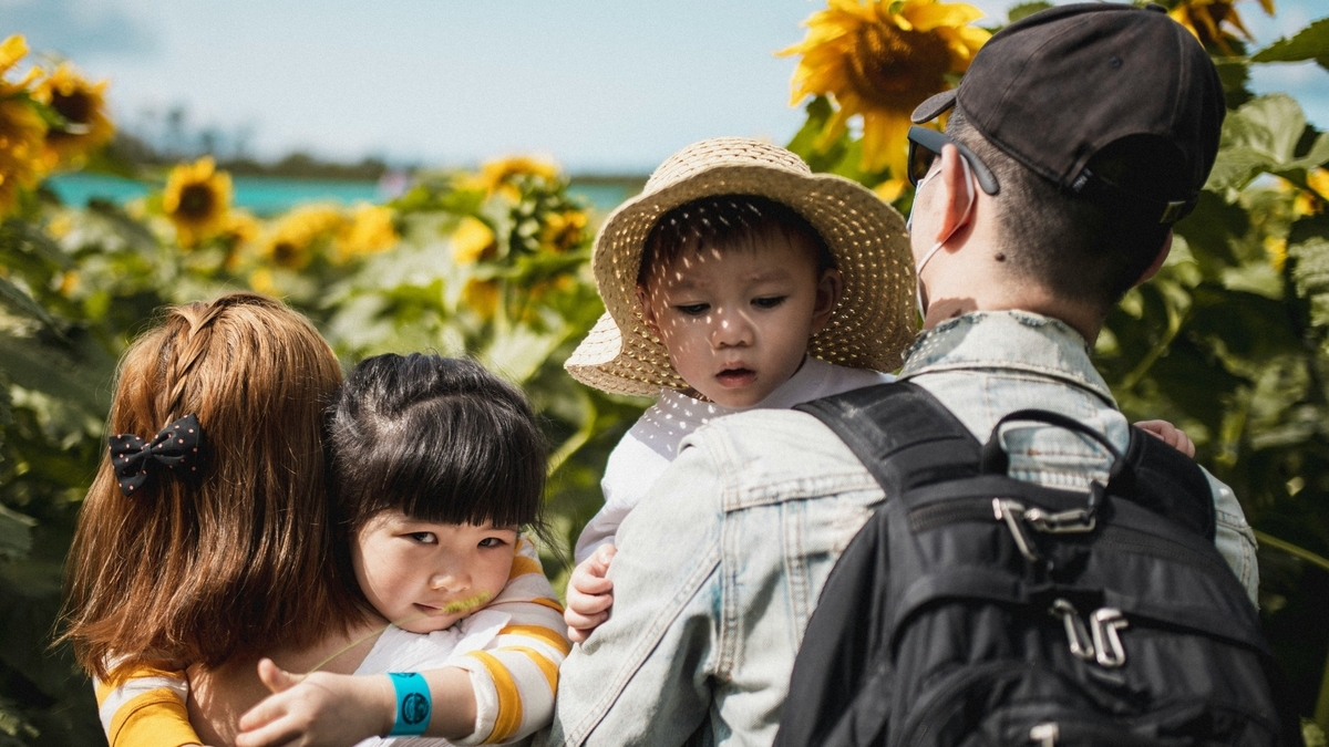 ひまわり畑の中で、帽子をかぶった2人の子供をそれぞれ抱きかかえている両親の後ろ姿と横顔