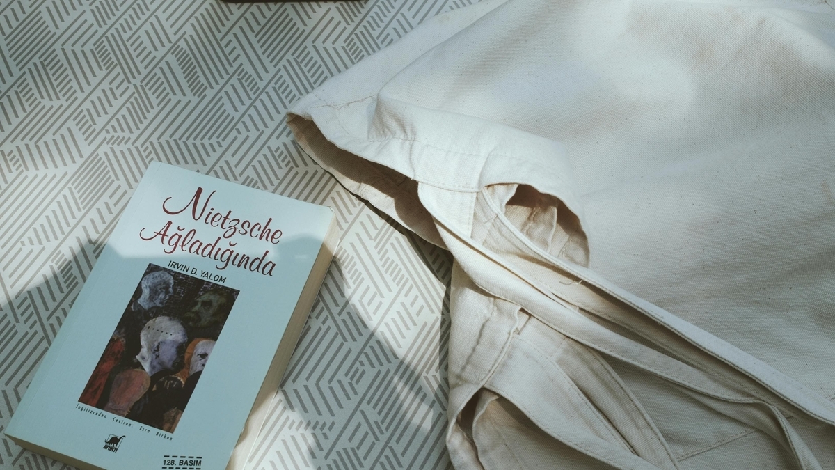 格子模様の敷物の上に、トルコ語の書籍「Nietzsche Ağladığında」と生成りの布バッグ、黒い電子機器が置かれている様子
