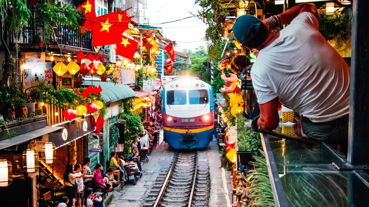 赤い国旗やランタンが飾られた狭い線路沿いの街並みと、そこを走る青と白の列車を、バルコニーからカメラで撮影している男性