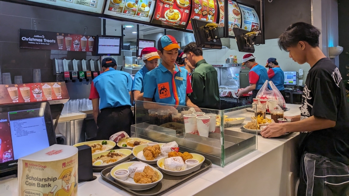 Jollibeeの注文カウンターの様子。青い制服と帽子を着用した複数の店員が調理や袋詰めをしており、カウンターの上にはフライドチキンやライスの乗ったトレイ、ドリンクカップ、募金箱が並んでいる