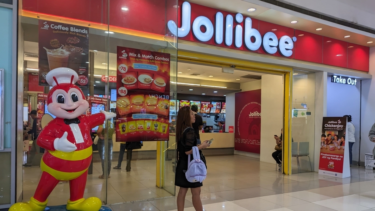 Jollibeeの店舗入り口。ガラス張りの外観に赤い大きなブランドロゴ看板があり、手前には赤い服を着た蜂のキャラクターの大きな置物が立っている。店内にはメニュー看板や利用客の姿が見える