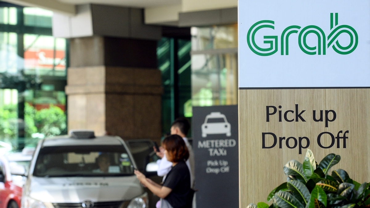 Grabのロゴマークが描かれた看板と、その奥に停車している配車サービス用の車をスマートフォンで撮影している利用客
