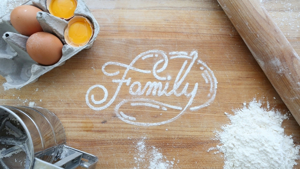 木製の作業台の上に広げられた小麦粉で「Family」の文字が描かれ、周囲に卵や麺棒、粉ふるいが配置されたベーキングの様子