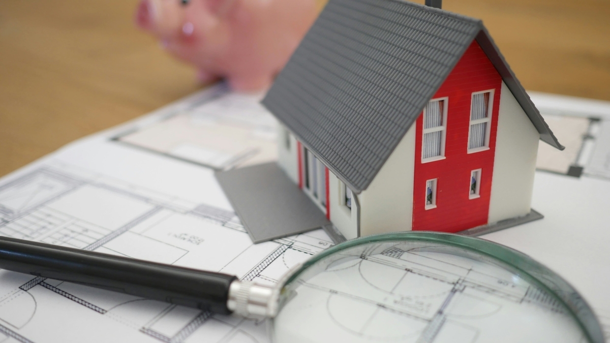 住宅の間取り図の上に置かれた、赤い壁とグレーの屋根を持つ一戸建ての模型と、手前に配置された虫眼鏡および奥のブタの貯金箱