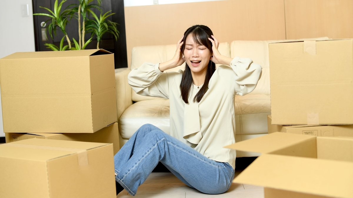 段ボール箱に囲まれた部屋の床に座り、両手を頭に当てて目を閉じ、困ったような表情や叫ぶような表情をしている女性