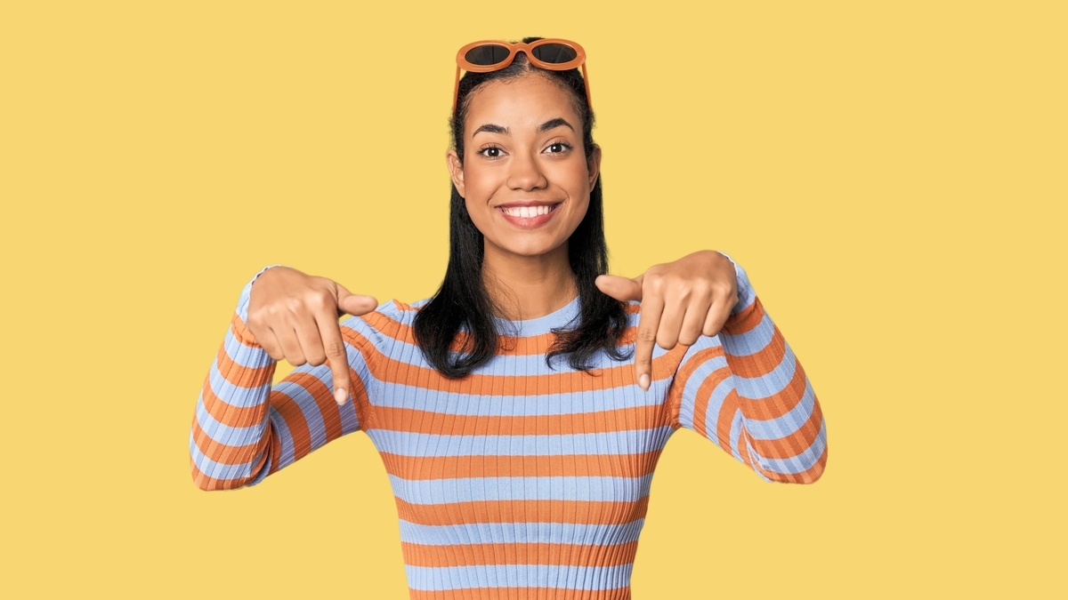 黄色の背景の前で、オレンジ色のサングラスを頭に乗せ、オレンジと水色のボーダー柄の服を着た笑顔の女性が、両手の人差し指で下を指さしている姿