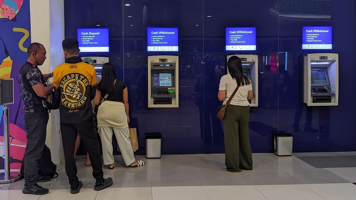 青い壁面に設置された4台のATMの前に、数人の男女が並んで現金を引き出そうとしている様子