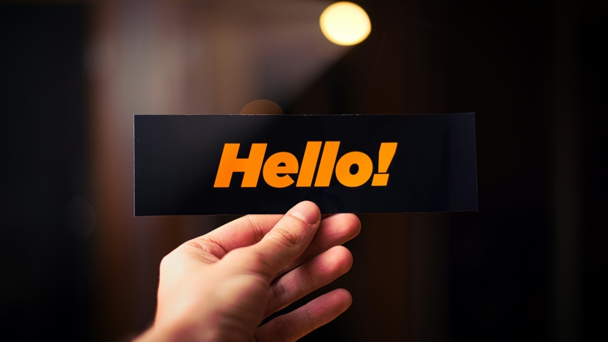 暗い背景の中で、オレンジ色の文字で「Hello!」と書かれた黒い長方形のカードを中央で指で挟んで持っている人の手