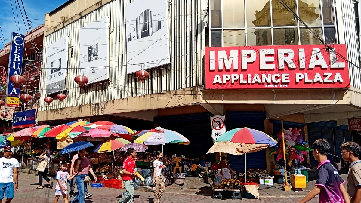 「IMPERIAL APPLIANCE PLAZA」の看板がある建物の前で、カラフルなパラソルや多くの人々が行き交う街頭風景