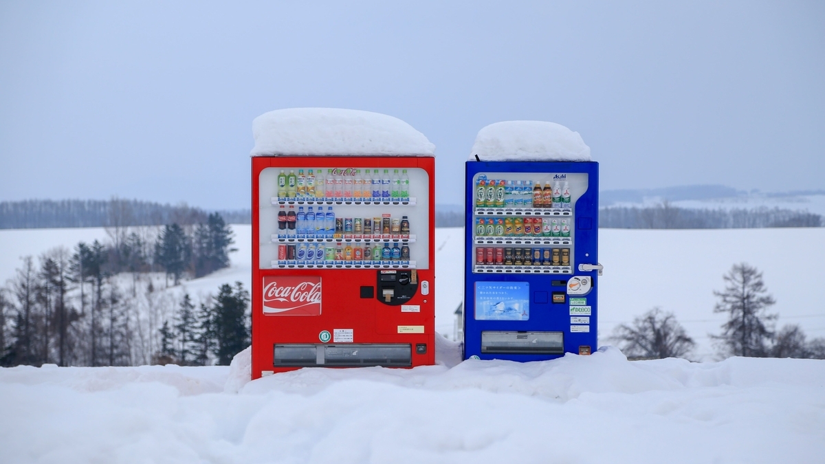 一面の雪景色の中に並んで設置された、雪がこんもりと積もった赤いコカ・コーラと青いアサヒの自動販売機