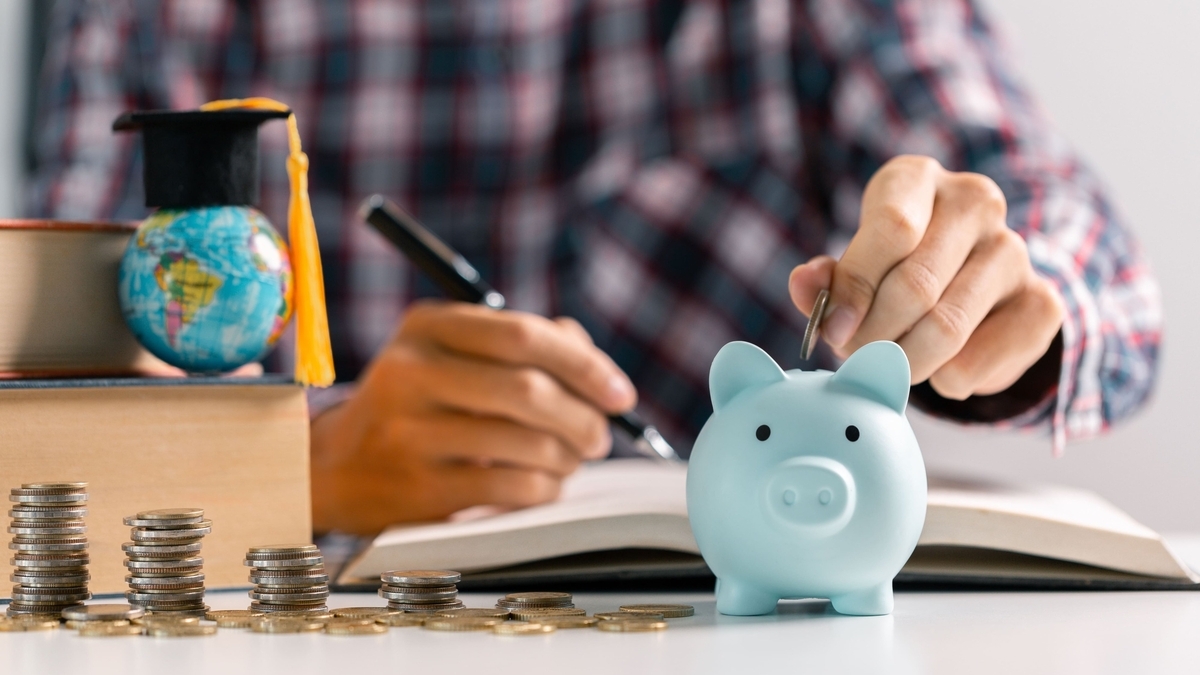 ノートにペンで書き込みをしている人の手前に、青い豚の貯金箱、積み上げられた硬貨、小さな卒業帽が乗った地球儀、本が置かれている様子
