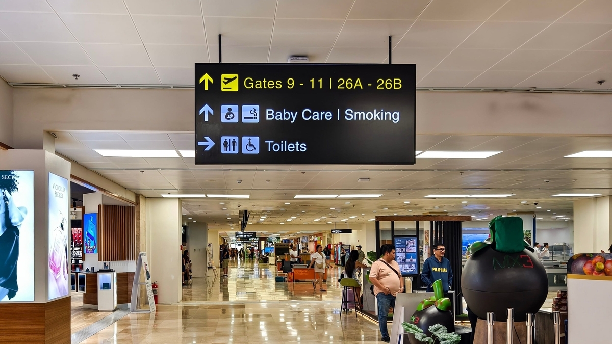 「Gates 9-11」や「Toilets」などの案内が表示された黒い電光掲示板が天井から吊り下がる、明るい照明に照らされた広々とした空港の搭乗ゲート付近の通路