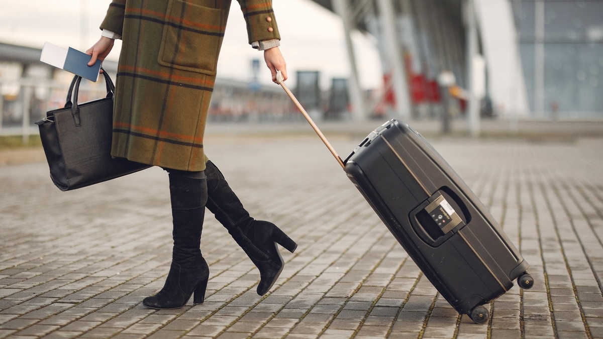 チェック柄のコートと黒いブーツを履いた人物が、片手にバッグと航空券を持ちながら黒いスーツケースを引いて歩く足元