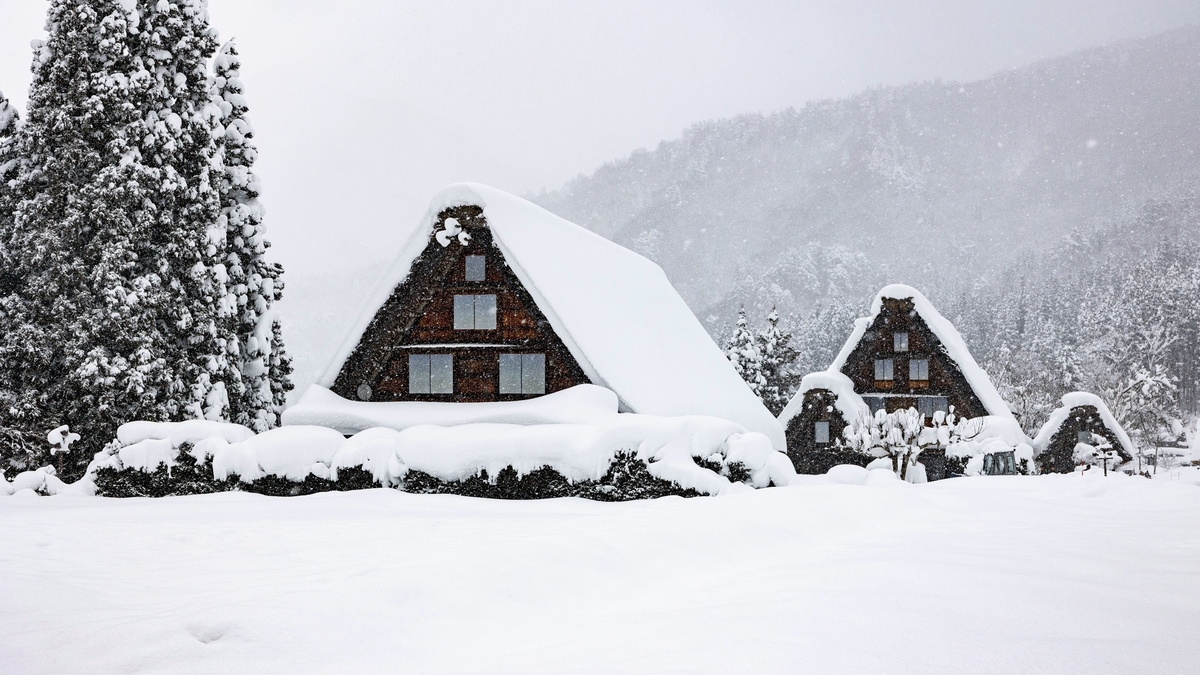 激しい雪が降る中、分厚い白雪に覆われた大きな三角屋根を持つ2棟の伝統的な木造家屋と、雪が積もった周囲の木々や山肌