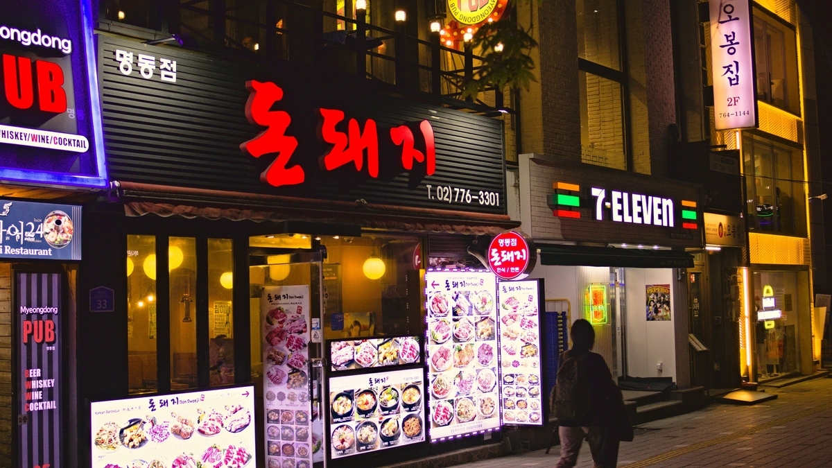 夜の街並みで赤く光るハングルの看板を掲げた飲食店と、その隣に並ぶセブンイレブンの店舗外観