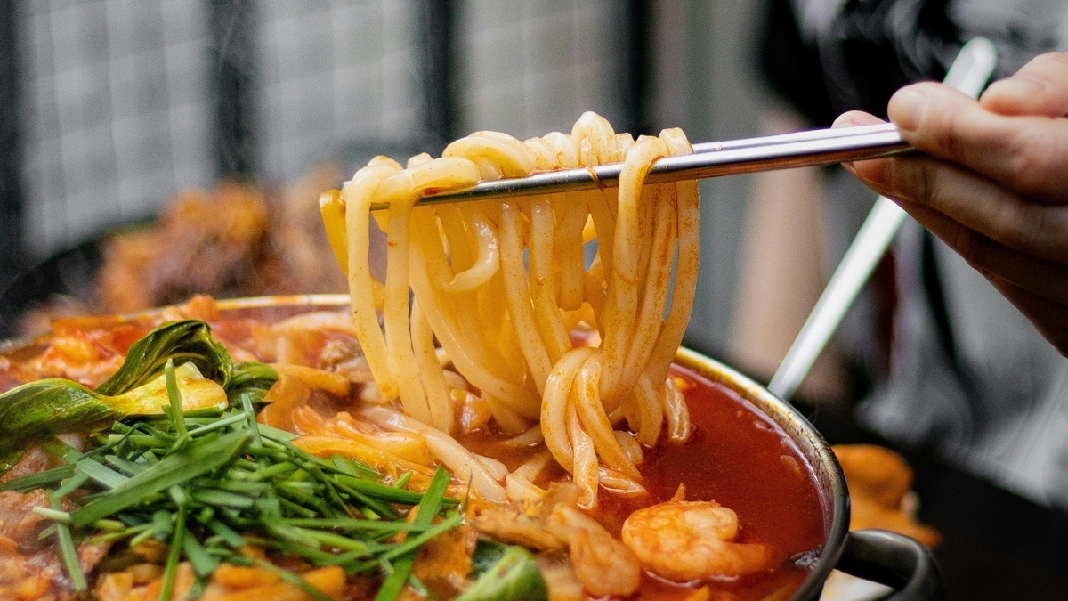 箸で持ち上げられた太い麺と、手前にニラやエビが添えられた赤く辛そうなスープの鍋料理