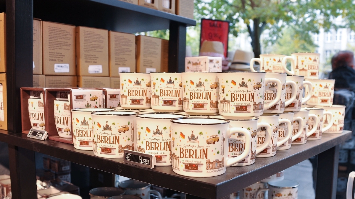 黒い棚に並ぶ、「Greetings from BERLIN」と文字が書かれた白いマグカップ。イラストが描かれたカップが手前や奥に多数重ねられている