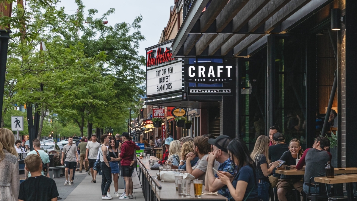 「CRAFT」と書かれた看板がある屋外の飲食店テラス席で、多くの人々がカウンターに座って飲み物を手に持ちながら談笑している賑やかな街の風景