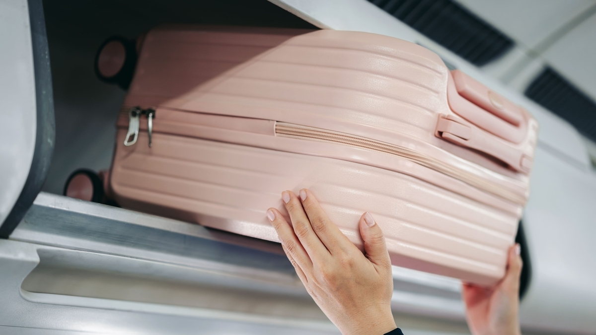 飛行機の座席上部にある白い荷物棚へ、両手を使って薄いピンク色のスーツケースを押し入れて収納しようとしている人の手元のクローズアップ