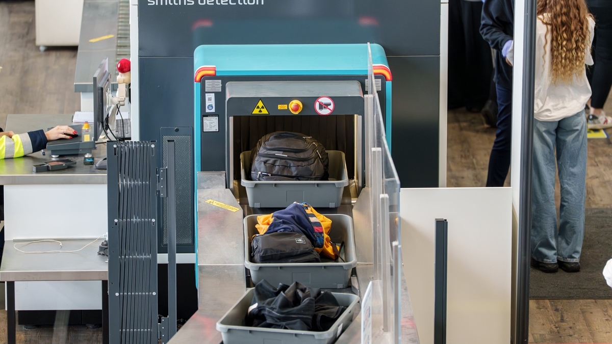 空港の手荷物検査場で、黒いリュックサックや衣類が載せられたグレーのプラスチックトレーがベルトコンベアに乗ってX線検査機へと進む様子