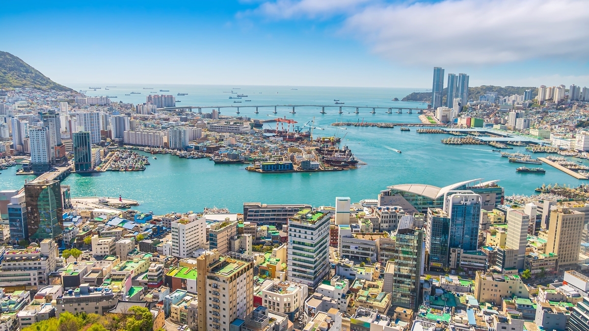 多くの高層ビルや住宅が密集する湾岸都市の全景。穏やかな青い海には多数の船が浮かび、奥には対岸を結ぶ長い橋が架かっている
