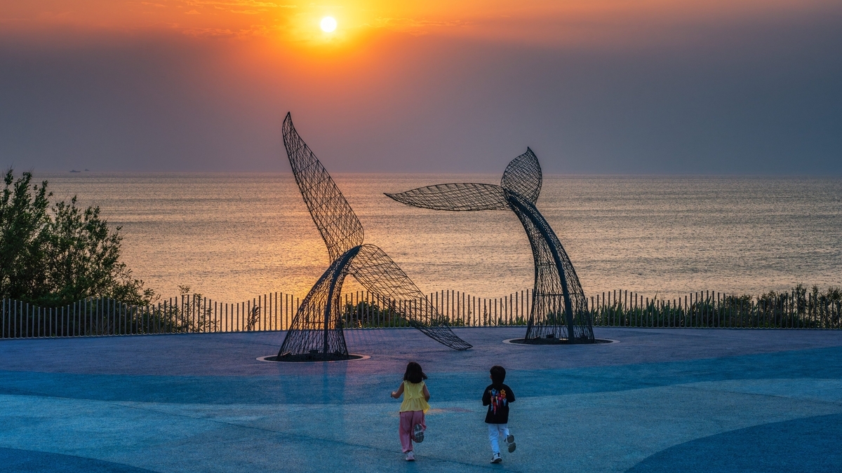 夕日が海をオレンジ色に染める中、並んで前方に走る2人の子どもの後ろ姿。その先にはクジラの尾びれを模した2つの大きなワイヤーアートがある
