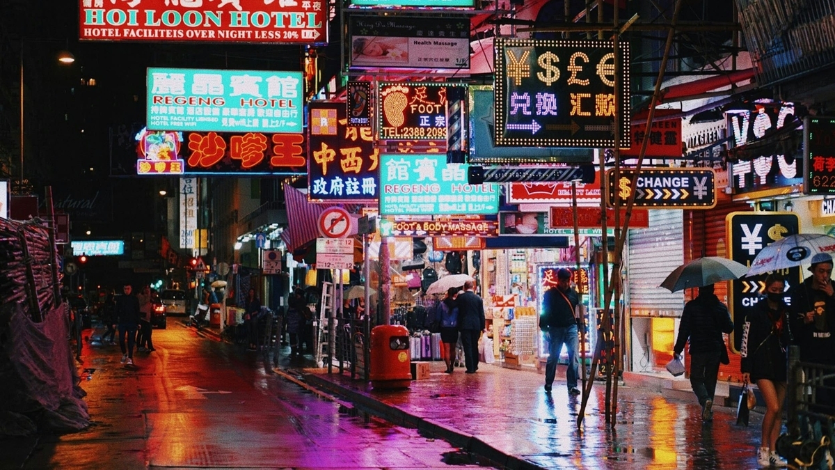 漢字や英語のネオン看板が多数きらめく夜の街並み。雨上がりの濡れた路面に光が反射する中、傘を差した人々が歩道を歩いている