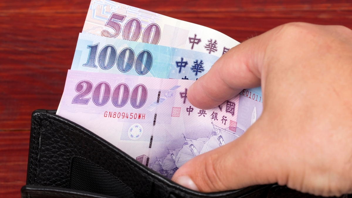 黒い財布から、紫色の2000元札、青色の1000元札、茶色の500元札の3種類の台湾元紙幣を右手で取り出している手元のクローズアップ。背景には赤茶色の木目の壁が映っています