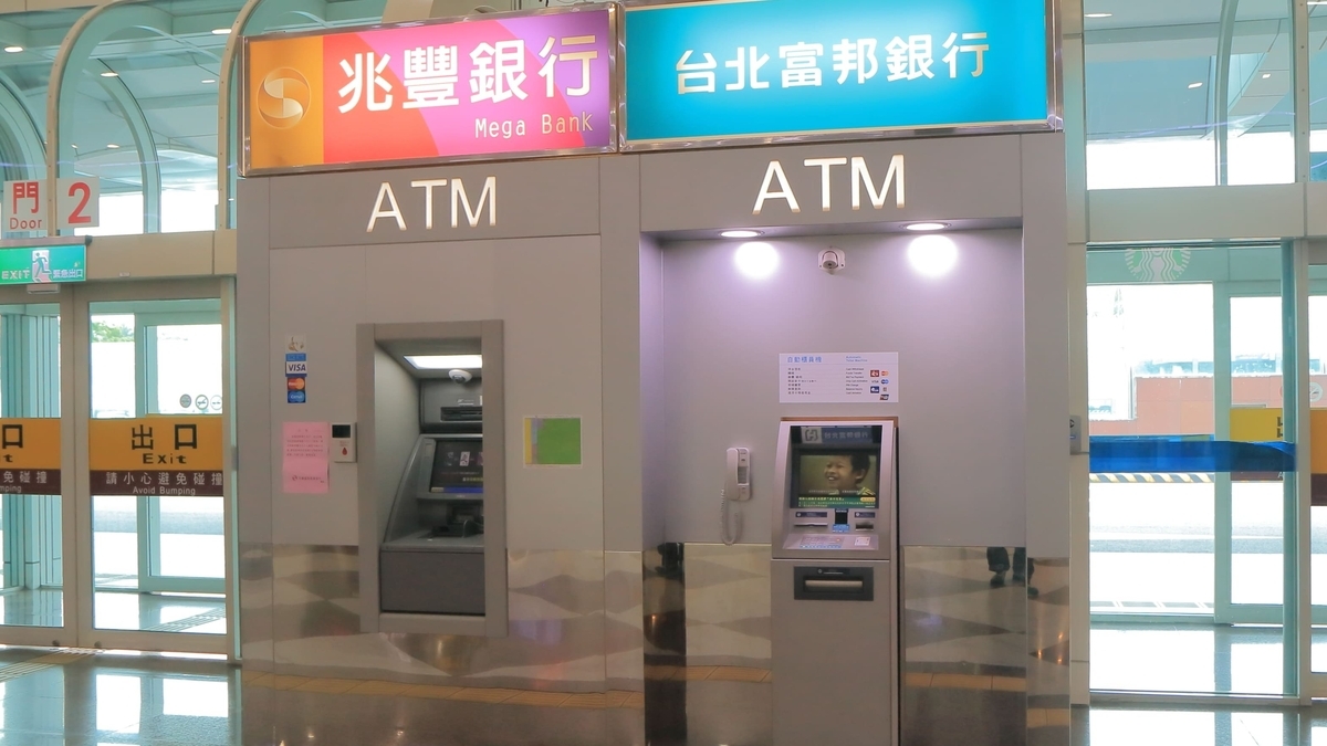 「兆豊銀行」と「台北富邦銀行」の看板が並ぶ屋内のATMコーナー。左側は壁に埋め込まれ、右側は独立した機械が設置されています。左奥には「出口 Exit」と書かれたガラス張りの扉が見えます。