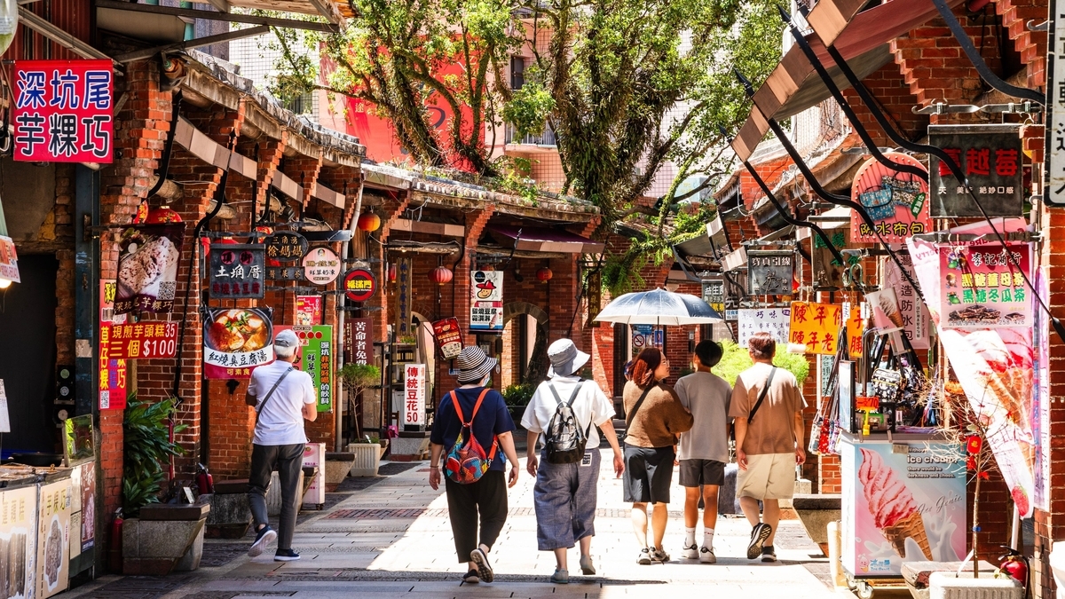 赤レンガ造りのレトロな建物と漢字の看板が並ぶ通りを、カジュアルな服装の観光客たちが奥へ向かって歩いている様子。通路の上には大きな木々が茂り、木漏れ日が地面を照らしています