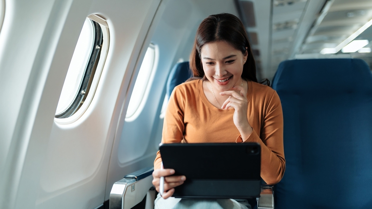 飛行機の窓際の座席に座り、タッチペンを手に持ちながらタブレット端末の画面を見て微笑むオレンジ色の服を着た女性