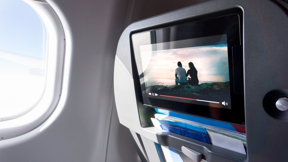 飛行機の窓際にある座席背面のモニターに、夕日を背に佇む男女の映像が映し出されている様子