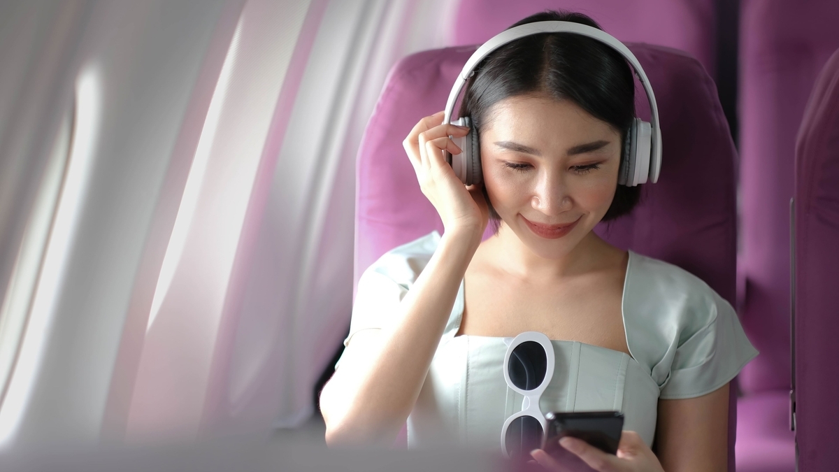 飛行機の機内で、白いヘッドホンを装着しスマートフォンを操作しながら微笑む、紫色の座席に座った女性
