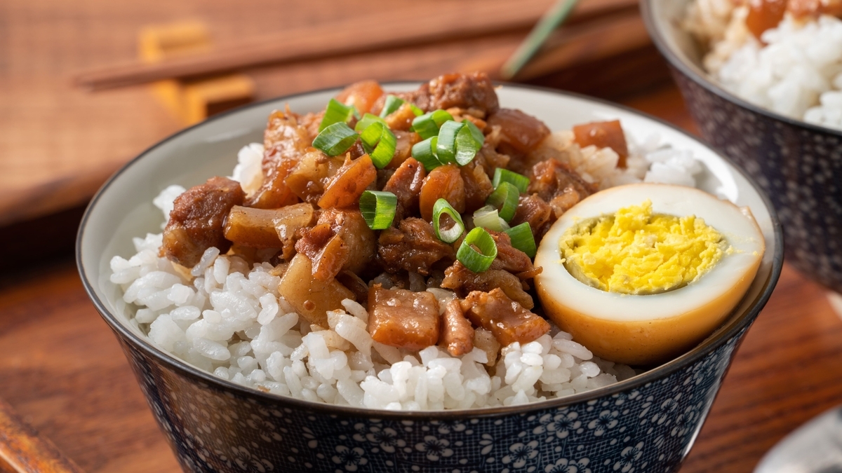 青い花柄の器に盛られた白米の上に、甘辛く煮込まれた刻み豚肉と刻み青ネギがトッピングされ、半分にカットされた味付け卵が添えられたルーローハンのクローズアップ