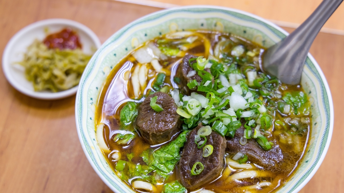 緑色の模様が入った白い器に盛られた、濃い茶色のスープに中太の麺と大ぶりの牛肉の塊、刻んだ青ネギと白ネギがトッピングされた牛肉麺のクローズアップ