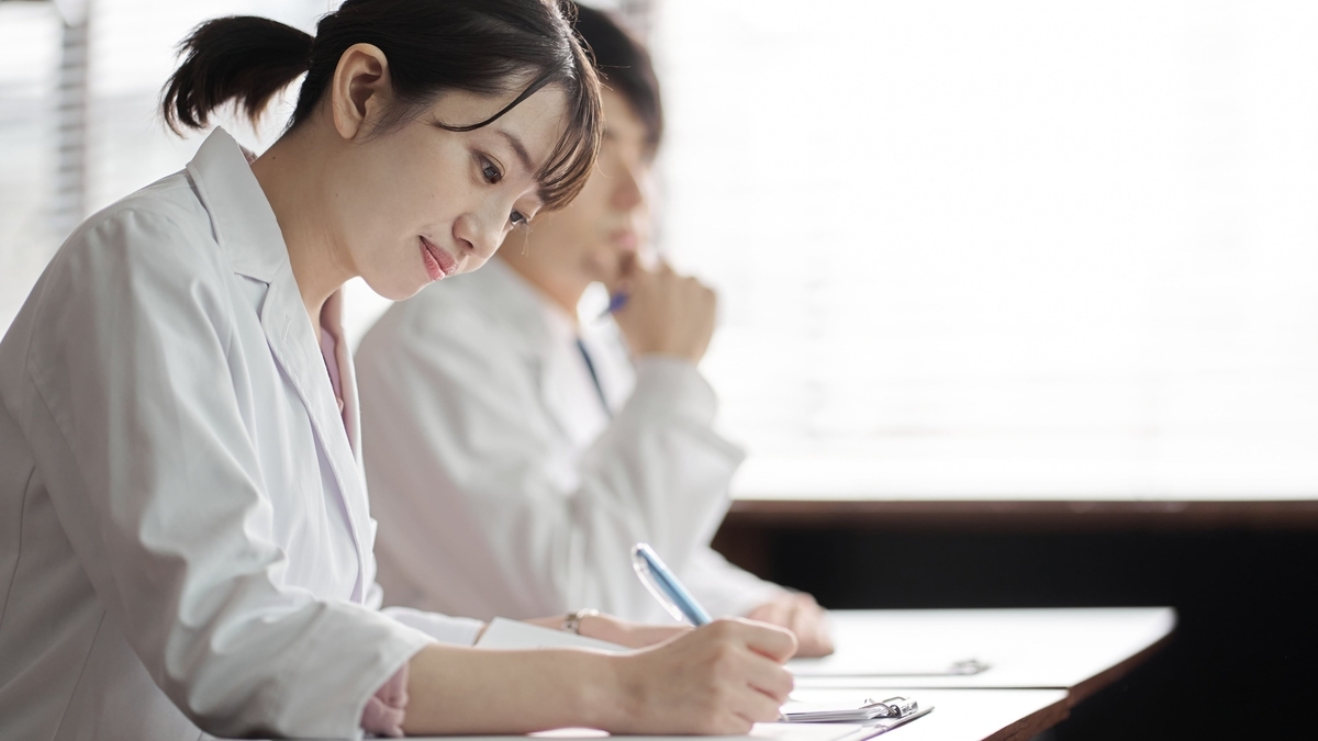白衣を着た髪を後ろで結んだ女性が、手元のクリップボードにペンで書き込みをしている。その背後では、同じく白衣を着用した男性が顎に手を当てて前方を見つめている