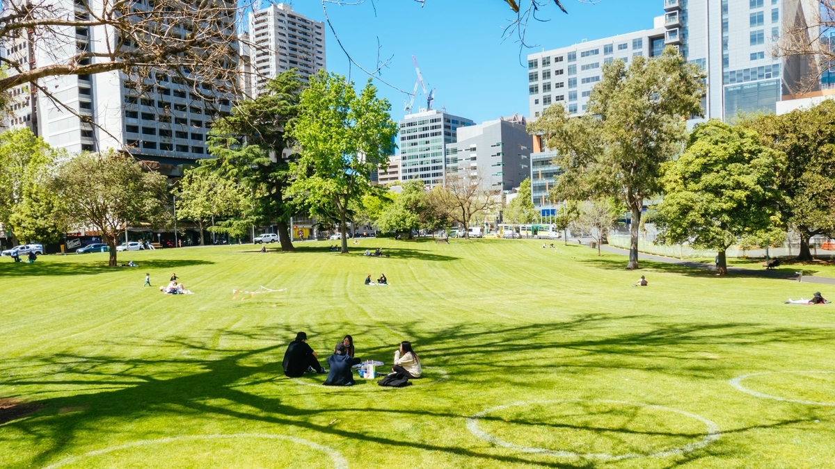 青空のもと、高層ビル群に囲まれた広い芝生の公園で、木陰に座りながらピクニックや休憩を楽しむ複数の人々